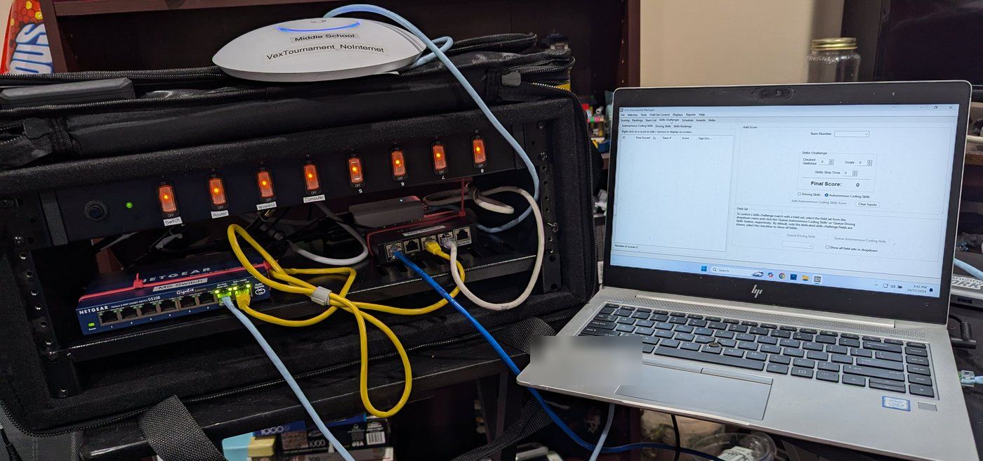 An open rack bag with a power strip of labelled switches, a network switch and a router, the Wi-Fi access point resting on top, and the Tournament Manager laptop beside it