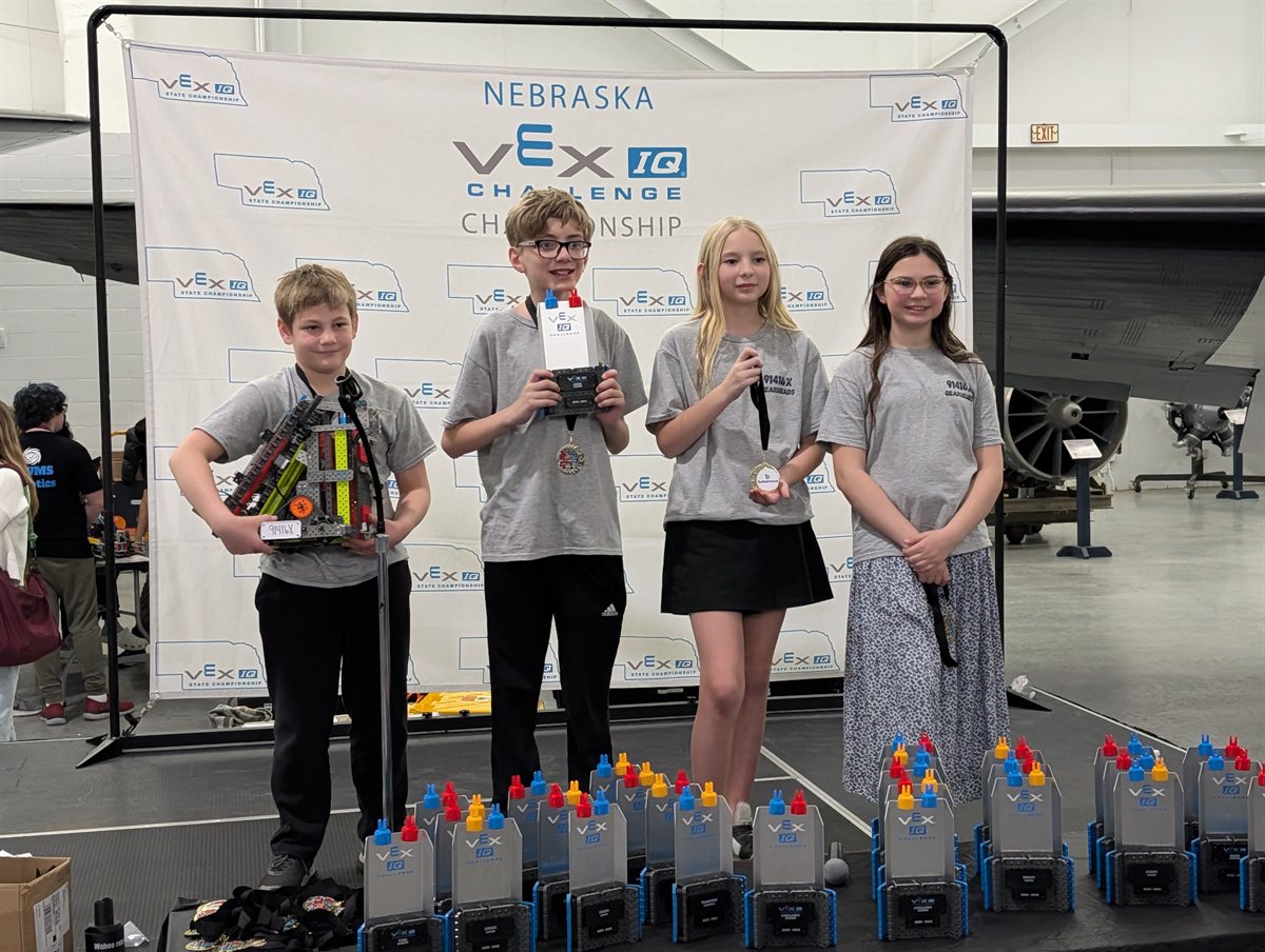 Four students from team 91416X Gearheads stand together in front of the Nebraska VEX IQ Challenge Championship banner, holding their robot, their controller and their medals.