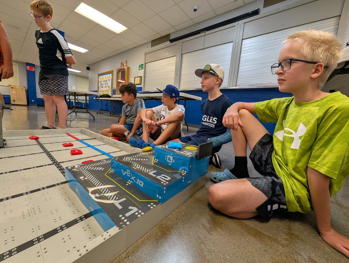 Students sitting on a gym floor around a VEX IQ Level Up field, with the tiered L1 and L2 goals and bean bags in the foreground