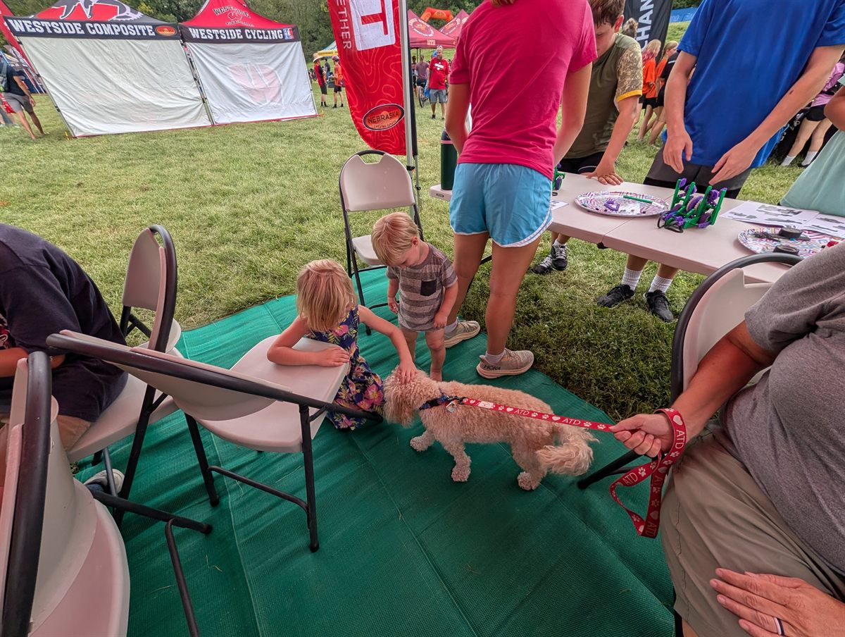 Two young children crouch to pet Bella, a light-coated therapy dog on a leash, with the robot building table just behind them