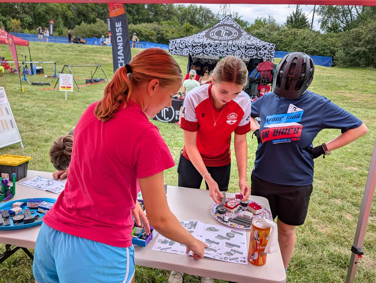 A girl in an Elkhorn Composite mountain bike race jersey assembles a VEX IQ robot while another rider, still wearing her helmet and a Nebraska NICA shirt, looks on