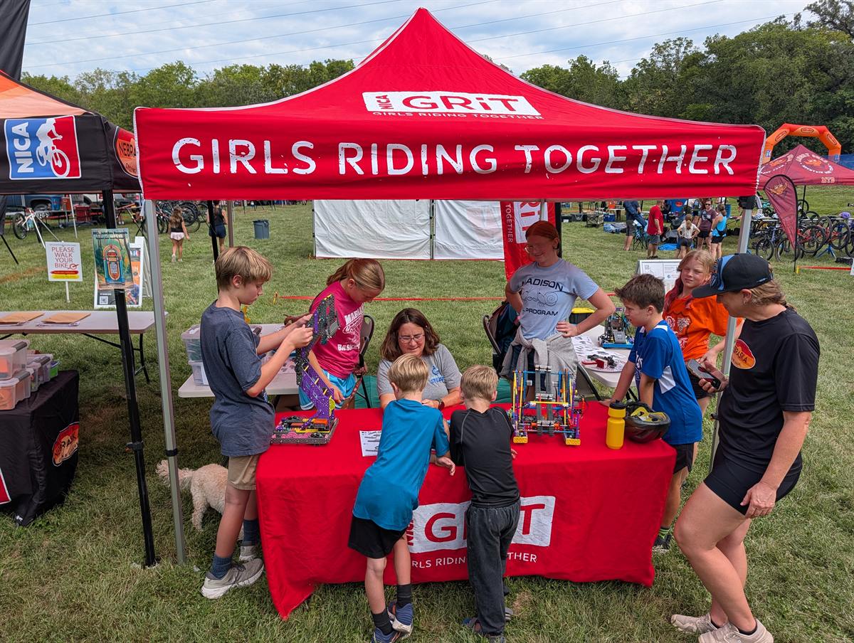 Kids gathered around a table of VEX IQ robots under a red Girls Riding Together tent at a mountain bike race
