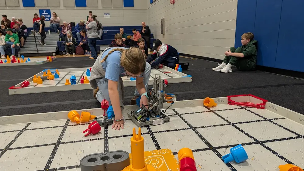 A student focused on programming their robot at a VEX IQ tournament field, with families watching from the stands