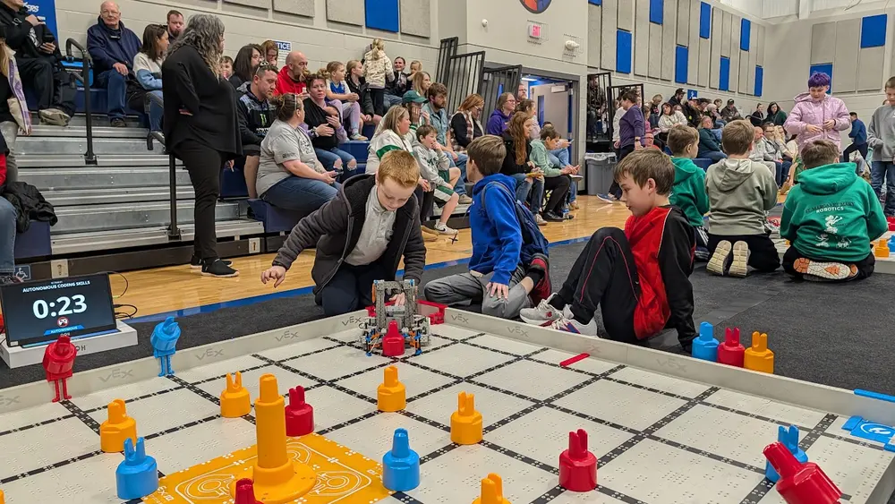 Students competing at a VEX IQ tournament field with the match scoreboard and families watching