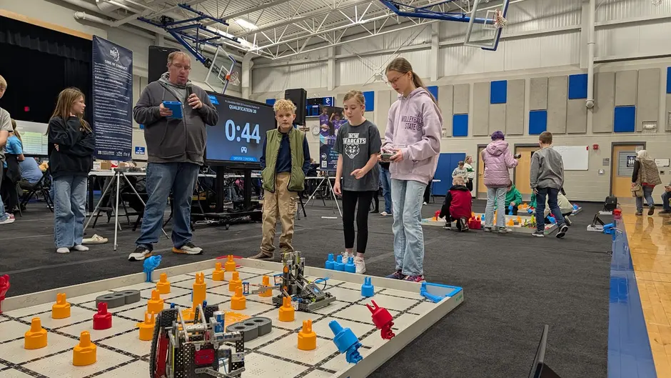 Students driving robots at a VEX IQ tournament field, with the match timer on the scoreboard, in a school gymnasium
